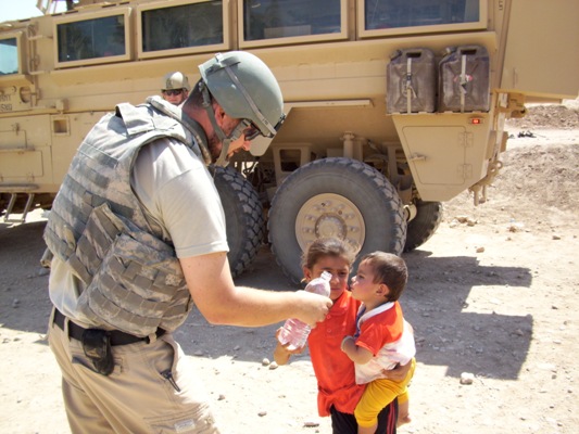 A serviceman handing drinking water to a woman carrying a young child beside an armoured vehicle